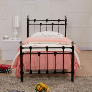 Single bed with black metal frame and polished chrome caps, dressed in light pink quilt and white pillow, with a white nightstand and lamp beside it.