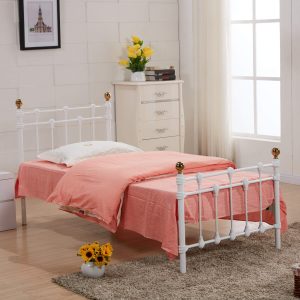 White metal single bed frame with polished gold caps, pink bedspread, white chest of drawers with yellow flowers, large window with sheer curtains, and beige rug with sunflowers.